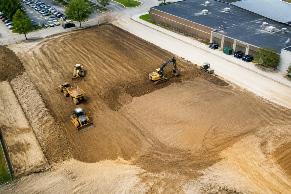 Aerial view of a commercial property site with heavy excavation equipment actively grading and preparing the foundation area, showing compacted soil, cleared land, and machinery positioned for concrete installation