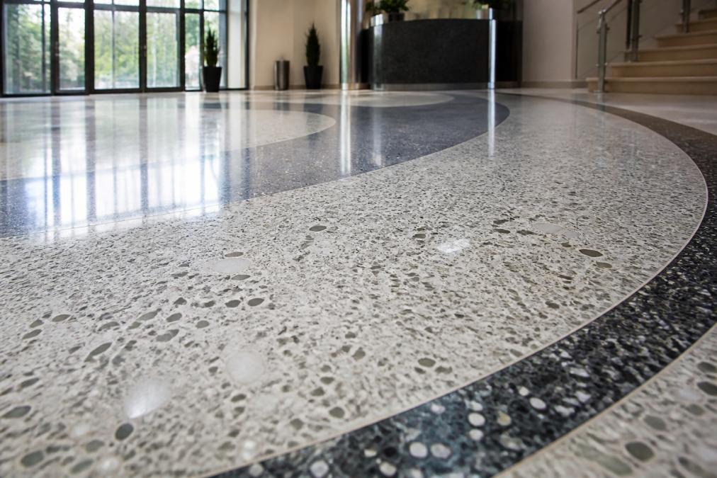 Close-up of polished terrazzo flooring with marble and glass aggregate in a modern commercial lobby, showing reflective surface, decorative inlay patterns, and professional finish under bright overhead lighting