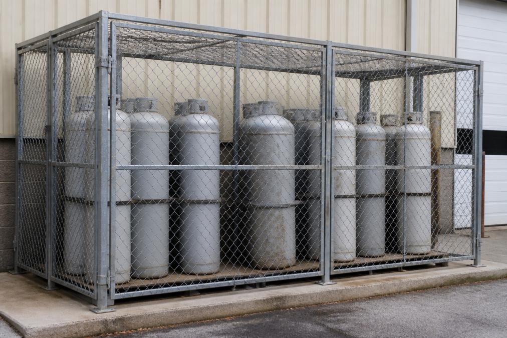 Secured galvanized cylinder storage cage positioned outdoors on an industrial property with multiple propane tanks organized vertically and safely spaced apart, showing proper ventilation clearance