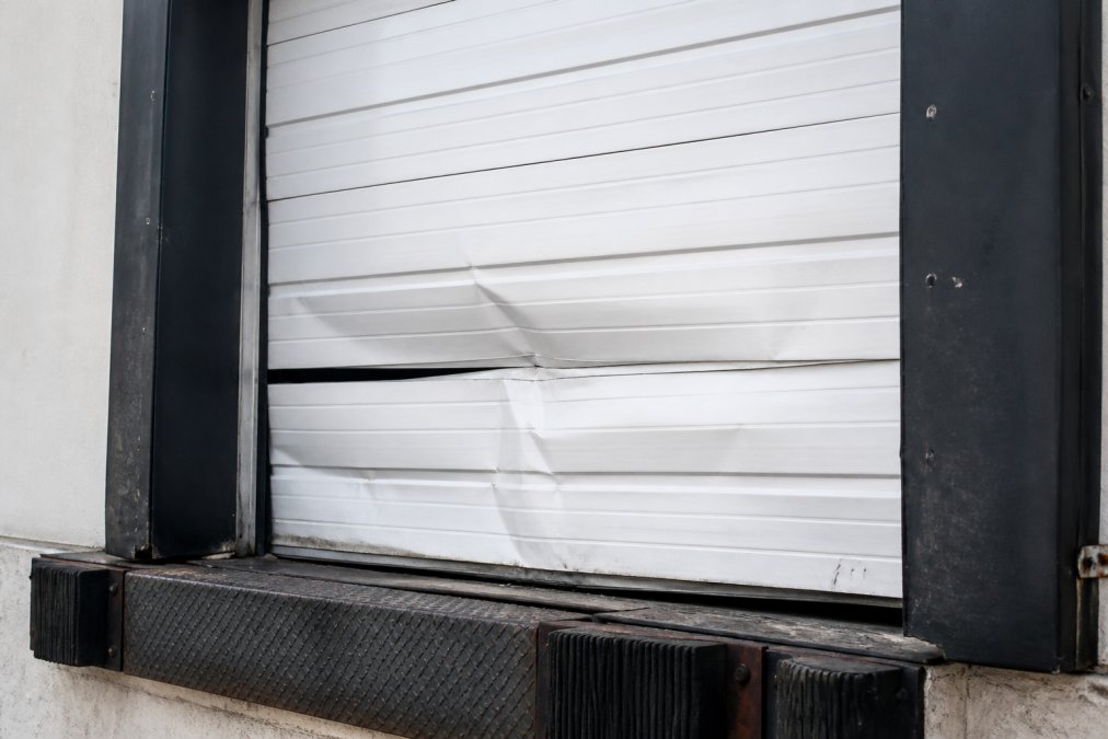Close-up of a damaged commercial garage door panel showing visible dents, bent metal, and frame misalignment on a warehouse loading dock door