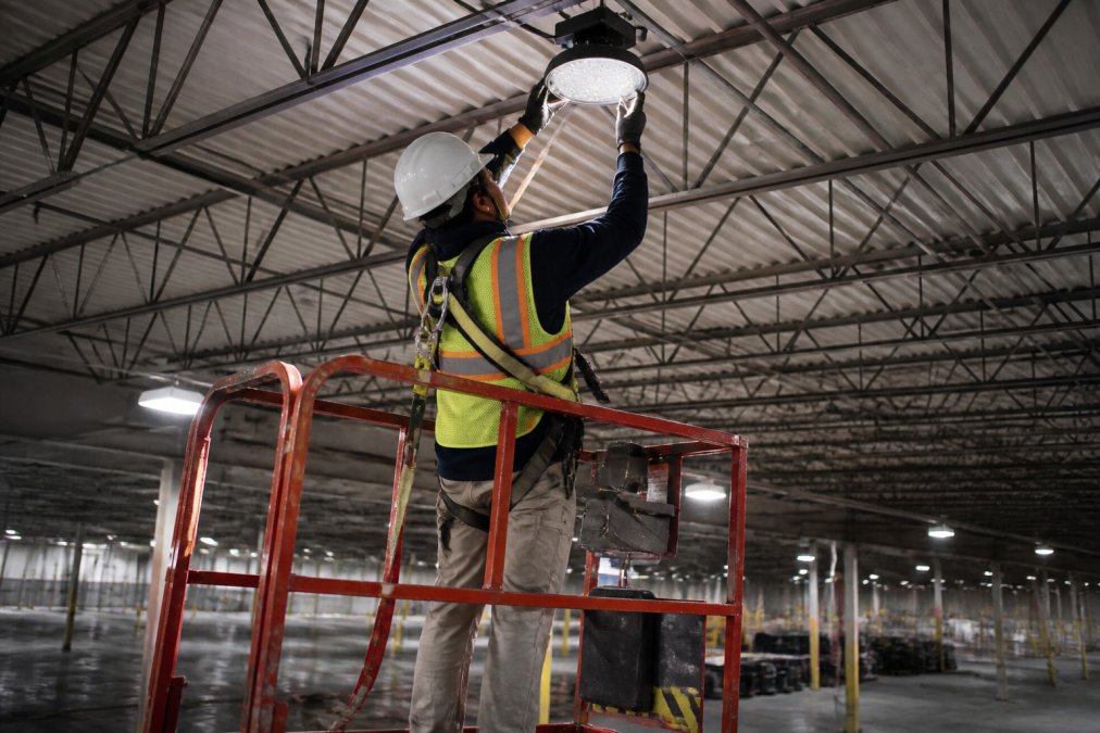 Certified technician installing LED fixture in high-bay warehouse ceiling using safety equipment and lift equipment, industrial facility with metal beams and concrete floor in background