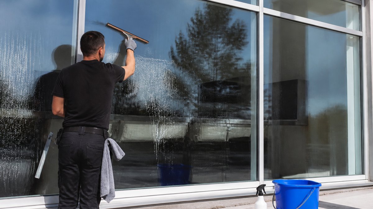A professional cleaner using a squeegee on a large commercial office window, with a bucket and cleaning supplies visible, bright daylight streaming through the glass
