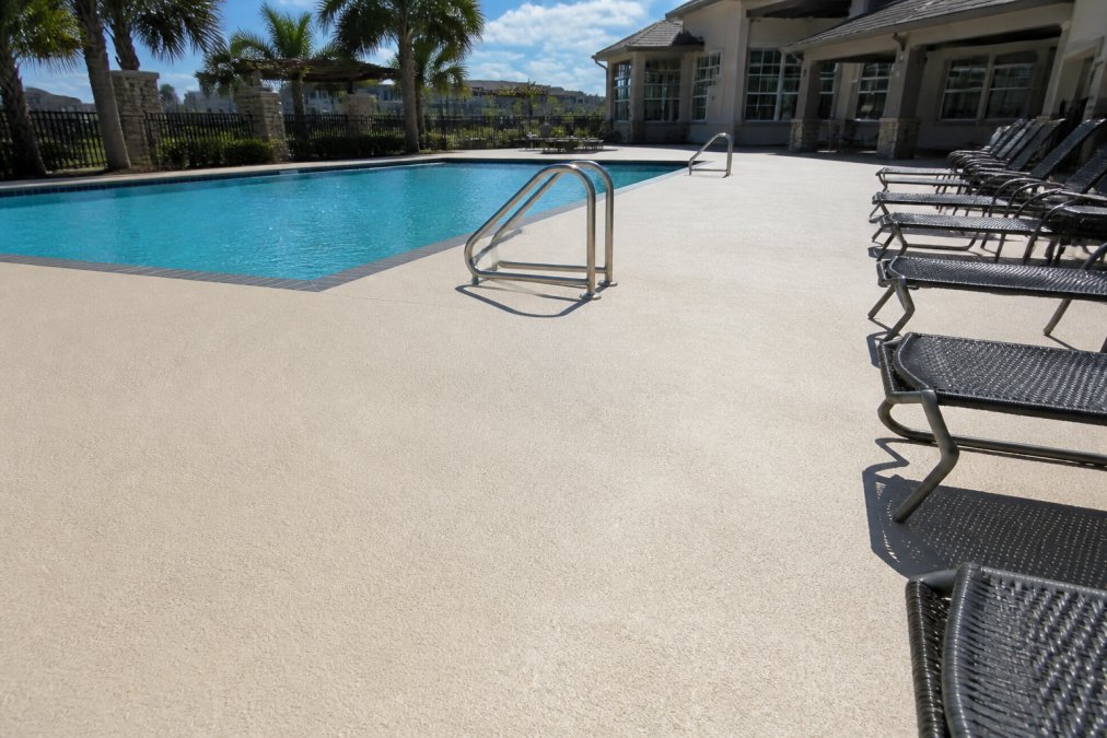 A freshly resurfaced commercial pool deck with clean, slip-resistant texture around a large outdoor pool, showing a smooth, professional finish under bright sunlight