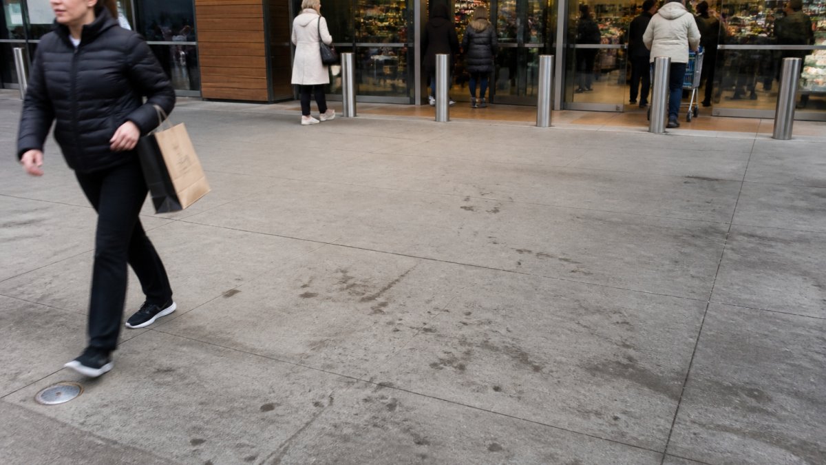Wide-angle view of busy commercial building entrance with multiple customers walking past, showing visible dirt marks and foot traffic stains on the concrete entrance area under natural daylight