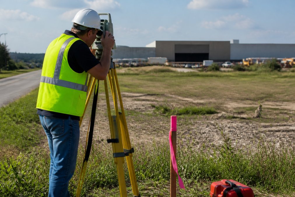 Professional land surveyor using surveying equipment and laser instruments on a commercial property, with property markers and measurement tools visible in the field