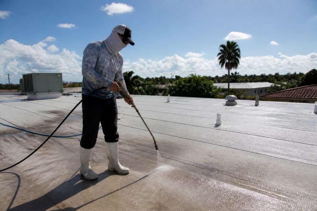Technician in safety gear applying soft wash solution to a commercial low-slope roof with a low-pressure spray wand under bright Florida sunlight