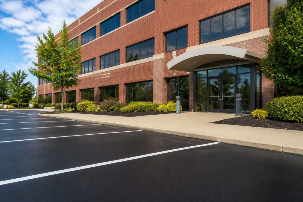A commercial office building with a clean, well-maintained brick facade, manicured landscaping beds with fresh mulch, a freshly sealed parking lot with clear striping, and polished entrance doors gleaming in afternoon sunlight