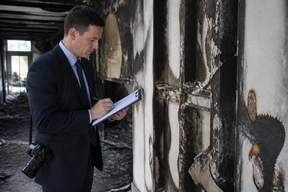 Professional insurance adjuster in business attire with clipboard and camera inspecting fire-damaged interior walls of commercial building, examining charred drywall and taking detailed notes