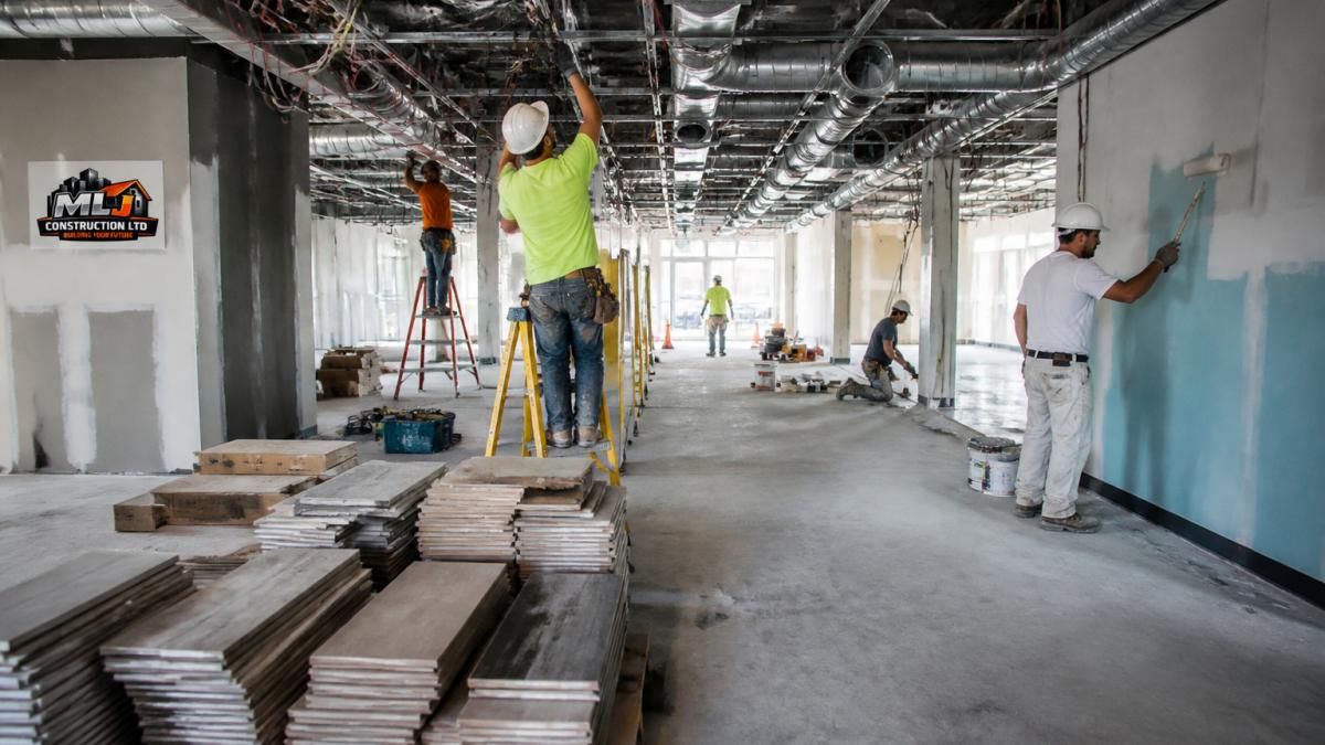 Interior of a commercial space under renovation showing exposed electrical wiring, HVAC ducts, various flooring materials stacked, and painters working on walls in different zones
