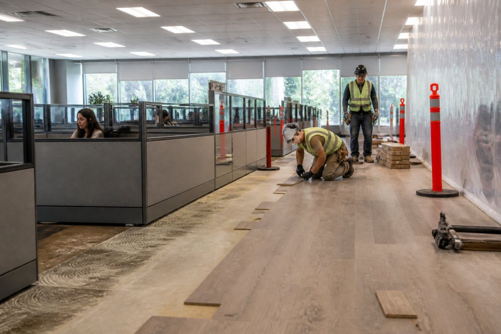 Professional construction workers installing new flooring in modern office space while existing cubicles with employees working remain visible in the background, demonstrating phased renovation work with proper safety barriers and natural daylight