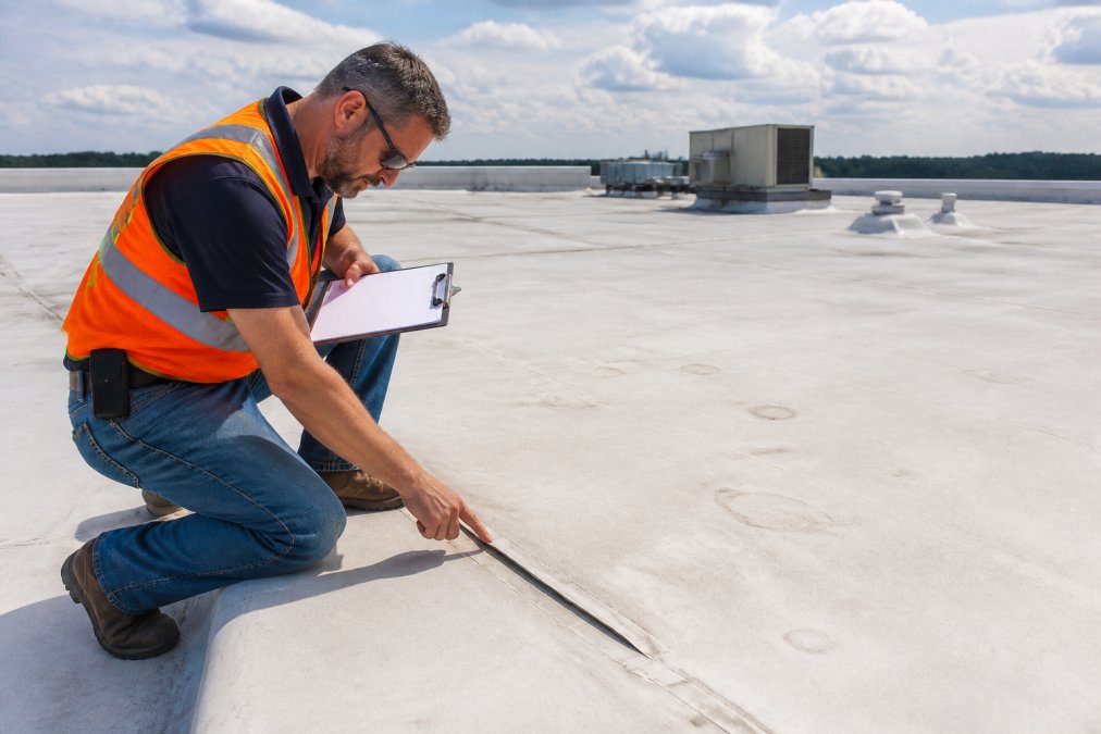 Professional inspector examining commercial roof membrane for blistering and seam separation, using clipboard to document findings, natural daylight across roofing surface