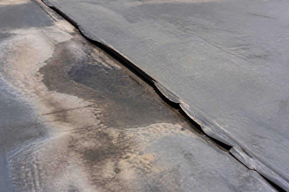 Close-up of deteriorated warehouse roof membrane showing visible seam separation, water staining, and UV-damaged material in natural daylight