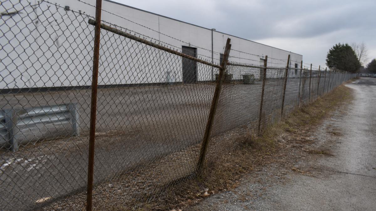 A commercial chain-link fence with visible rust and leaning posts along a warehouse perimeter, showing weather wear and physical damage on an overcast day