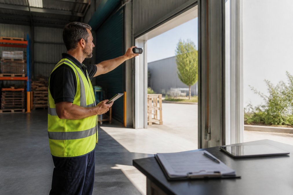 Security professional in high-visibility vest conducting on-site assessment at commercial warehouse entrance, photographing entry points and measuring distances with laser device, clipboard and tablet visible on nearby table, natural daylight streaming through loading bay doors