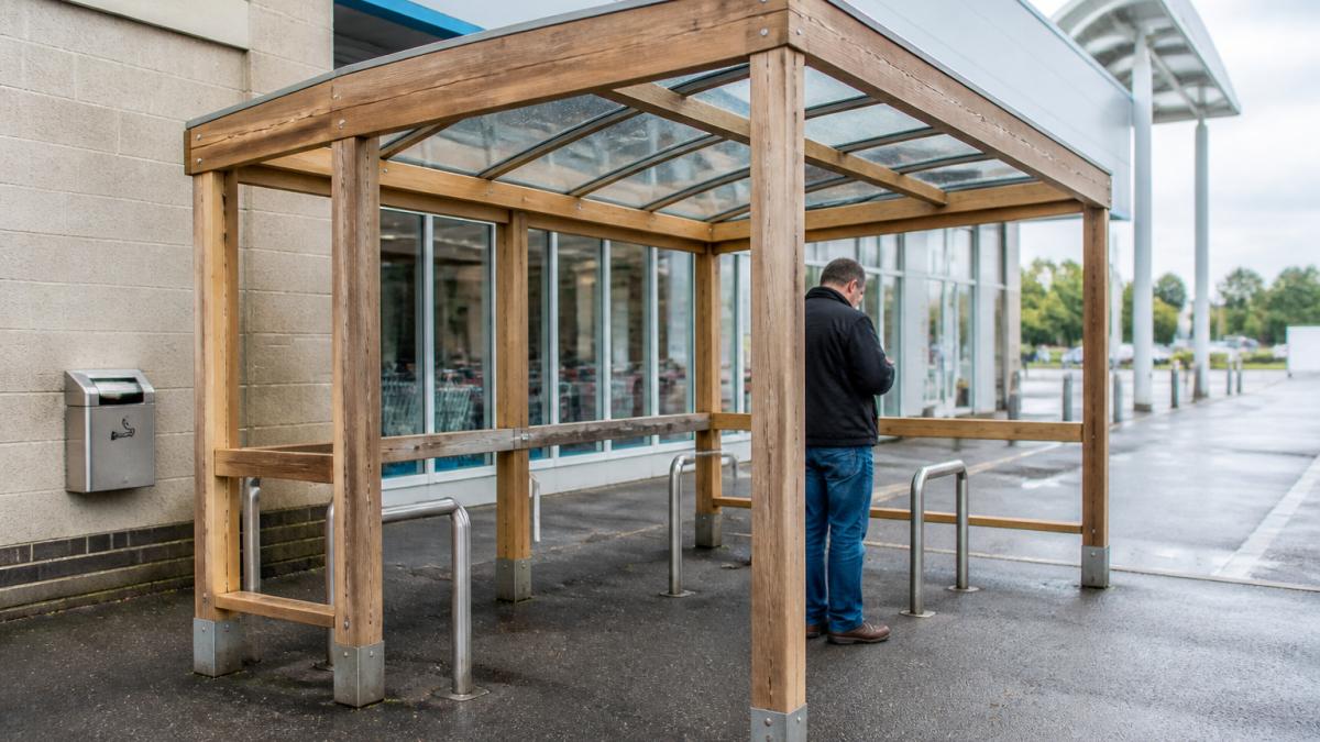 A modern open-sided smoking shelter with a glass roof and timber frame outside a retail building, with a person standing underneath and a wall-mounted cigarette bin nearby