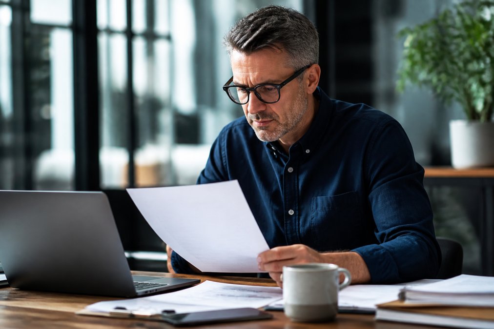 Business owner reviewing solar installation proposal and financial documents at a desk with a laptop and coffee, natural office lighting
