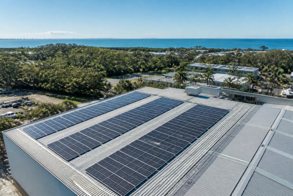 Aerial view of commercial building rooftop with multiple rows of solar panels installed, Sunshine Coast landscape and blue ocean visible in the background, bright midday sunlight