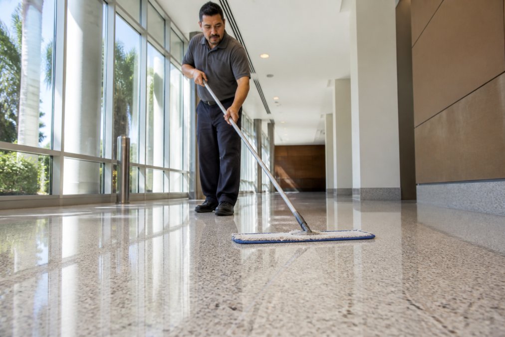 Facility maintenance worker using microfiber dust mop on polished terrazzo floor in bright commercial hallway with natural light, demonstrating proper technique