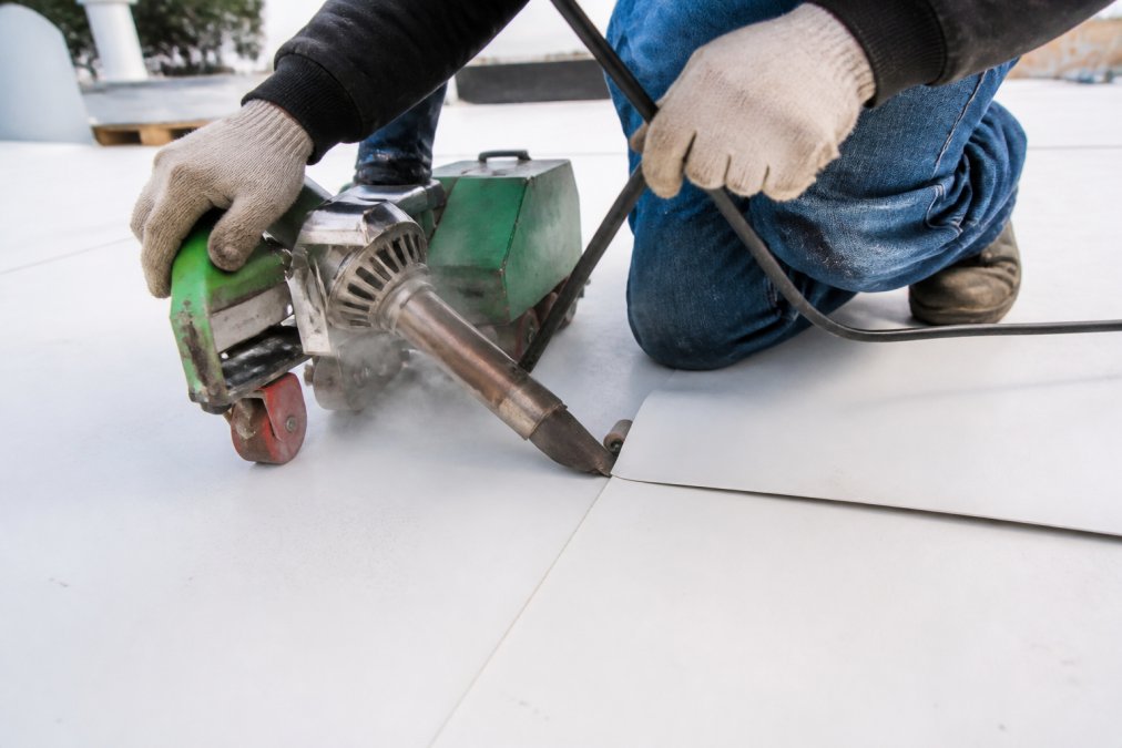 Close-up of a roofing technician in work gloves guiding an automatic hot-air welder along overlapping white TPO membrane seams on a commercial rooftop, with the welder nozzle glowing and steam visible at the weld zone