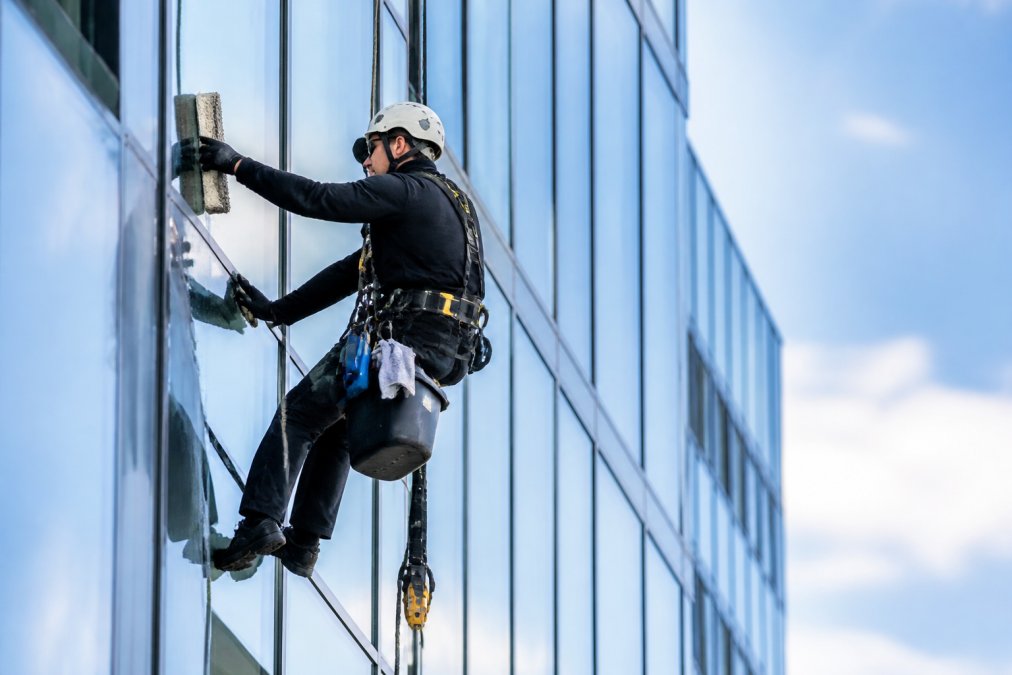 Professional window cleaner with safety harness secured to building exterior, demonstrating proper fall protection equipment setup on commercial high-rise facade