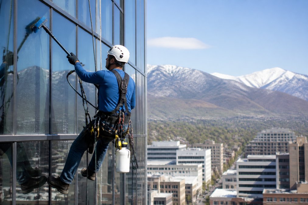 Professional window cleaning technician in safety harness and hard hat working on exterior of modern high-rise office building with water-fed pole system, demonstrating access difficulty and specialized equipment requirements