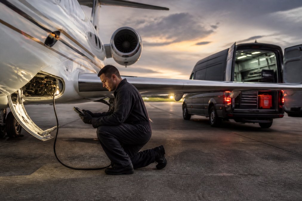 Aircraft maintenance technician in work coveralls kneeling beside a private jet on an airport tarmac at dusk, holding a diagnostic tablet connected to the avionics bay, with a mobile AOG response van parked nearby