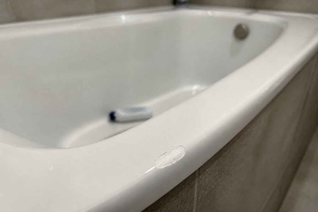 Close-up of a chipped white acrylic bathtub rim showing visible white damage against the glossy surface, with a dropped shampoo bottle nearby on the tub floor