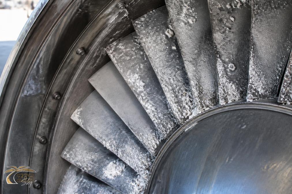 Close-up of jet engine compressor blade section showing salt deposits and white crystalline buildup on blade surfaces with visible surface pitting and corrosion damage in harsh coastal lighting