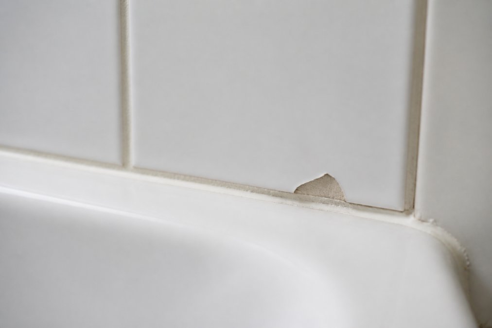 A close-up photograph of a chipped white bathroom tile edge next to a bath, with natural light showing the damaged glaze and surrounding grout
