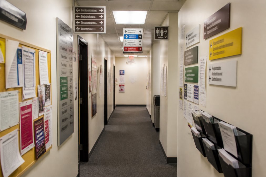 Cluttered office hallway with overlapping directional signs, multiple competing messages in different fonts and colors, and visual confusion from too much information displayed simultaneously on walls