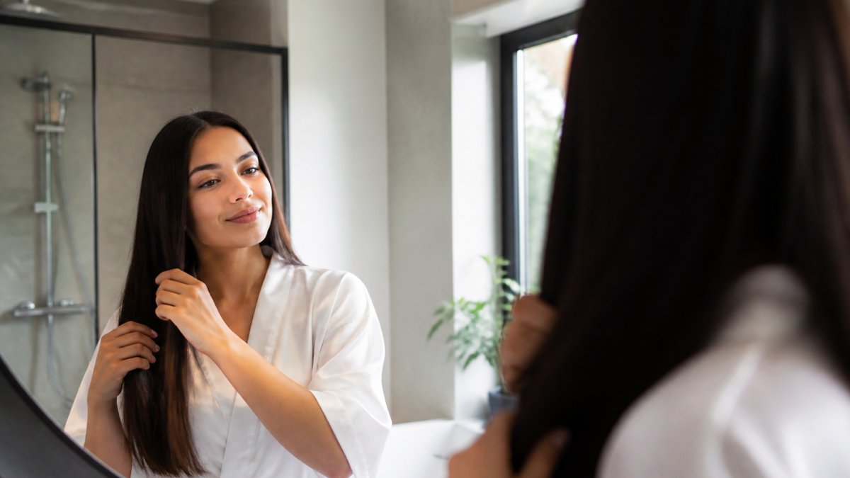 A woman with smooth, shiny hair looking at her reflection in a bathroom mirror, running her fingers through her hair with a satisfied expression, soft natural lighting