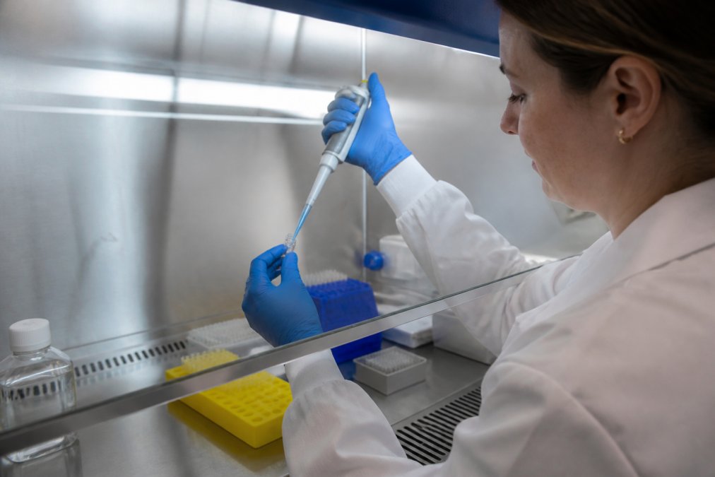Researcher in white lab coat carefully reconstituting a lyophilized peptide vial using sterile technique with a micropipette, wearing nitrile gloves and working under a biosafety hood with controlled lighting