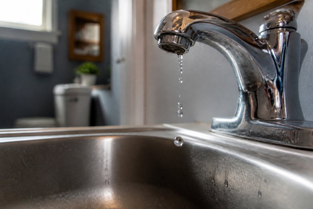 Close-up of water droplets falling from a leaking faucet into a stainless steel sink, with blurred background showing a residential bathroom