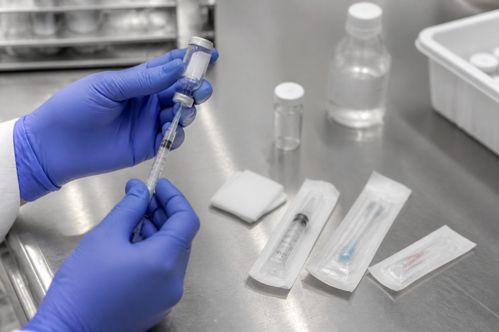 Close-up of a researcher's gloved hands preparing a subcutaneous injection with sterile supplies arranged on a clean stainless steel laboratory bench under bright overhead lighting