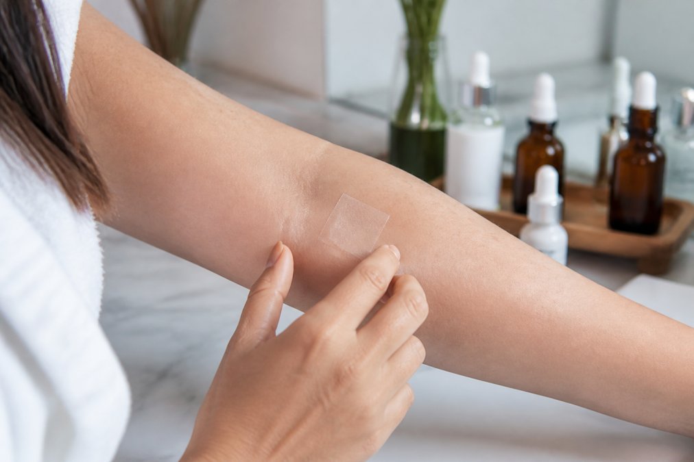 Close-up of a woman's forearm with a small square of clear patch test tape applied, soft natural bathroom lighting, skincare bottles blurred in background