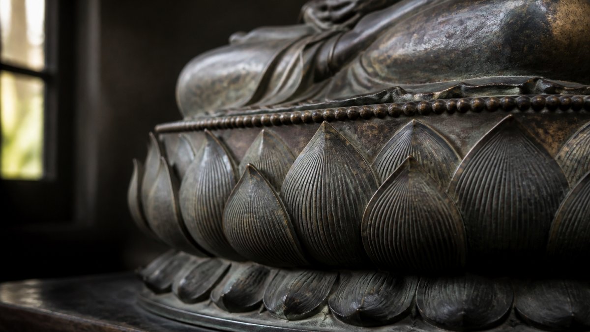Close-up photograph of an antique bronze Buddhist statue base carved with open lotus petals, soft natural window light, dark neutral background, shallow depth of field