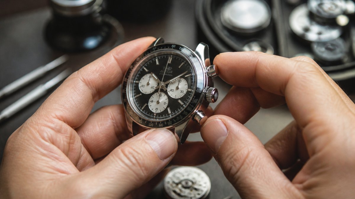 Professional watchmaker's hands holding a chronograph watch with polished steel case, showing subdials and pushers in sharp focus, with natural workshop lighting highlighting the tachymeter scale and chronograph mechanism