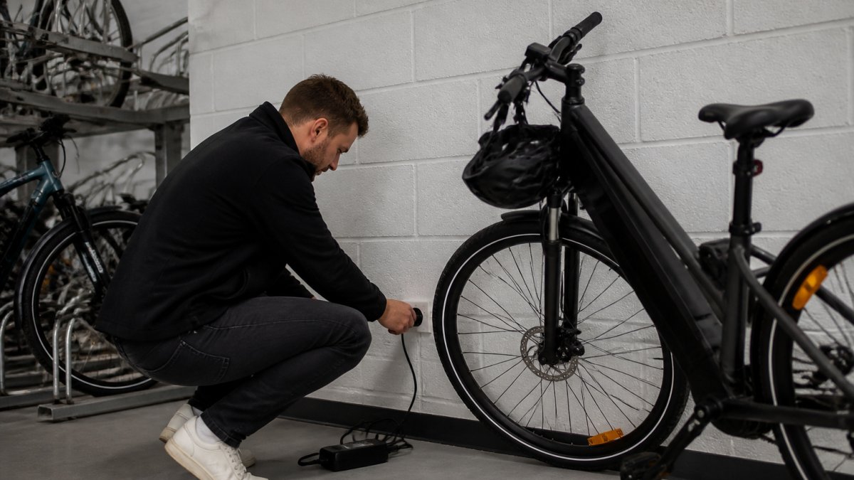 A commuter plugging an e-bike battery charger into a wall socket in a secure office bike store, with a parked e-bike leaning nearby