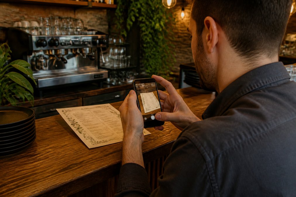 A restaurant owner sitting at a wooden counter with a smartphone, photographing a printed paper menu under warm café lighting, espresso machine visible in the background