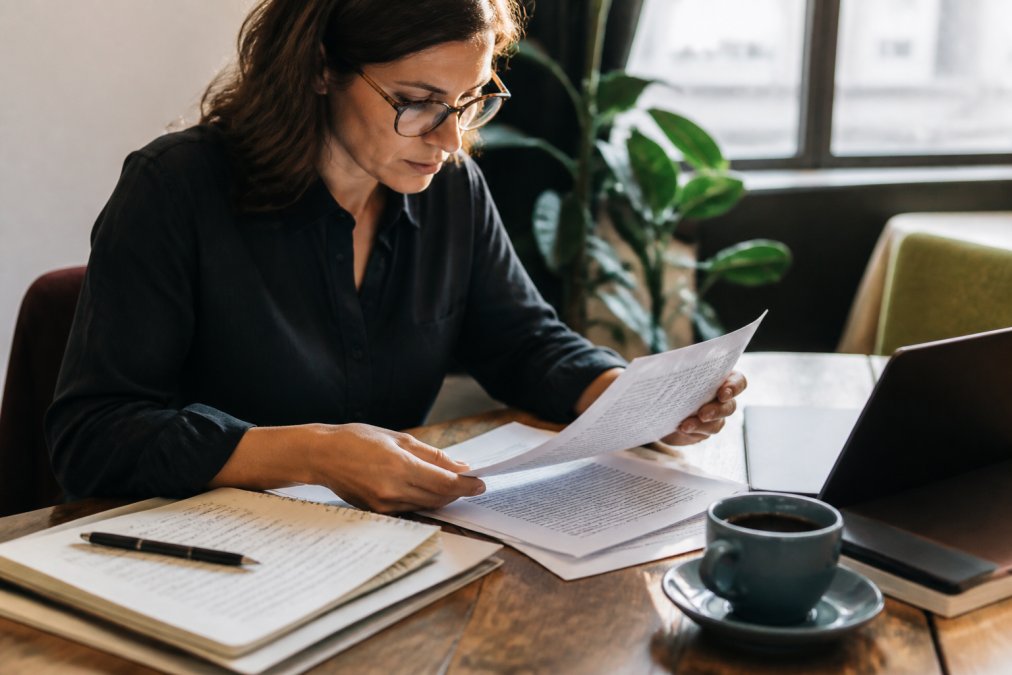 Professional reviewing document on laptop screen with glasses, coffee, and handwritten notes on work desk