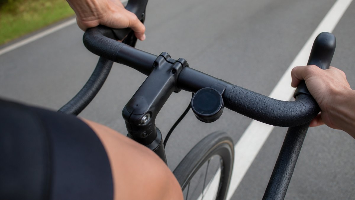 Close-up of a small circular bike navigation device mounted on the handlebars of a road bike, with a cyclist's hands resting on the brake hoods in the background on a paved road