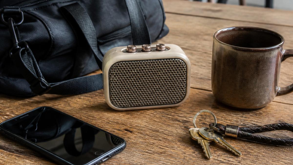 A compact retro-styled Bluetooth speaker sitting on a wooden desk next to a smartphone and a coffee mug, with a gym bag and keys nearby suggesting a commute or travel setup