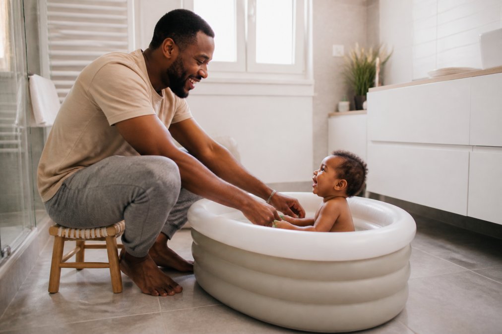 Un parent assis sur une chaise basse donnant le bain à son bébé dans une baignoire gonflable posée sur le sol d'une salle de bain lumineuse et moderne, le parent ne se penchant pas, souriant et regardant l'enfant