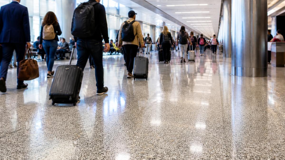 Busy airport terminal corridor with travelers rolling luggage, crowds walking, and polished flooring reflecting overhead lighting under high-traffic conditions