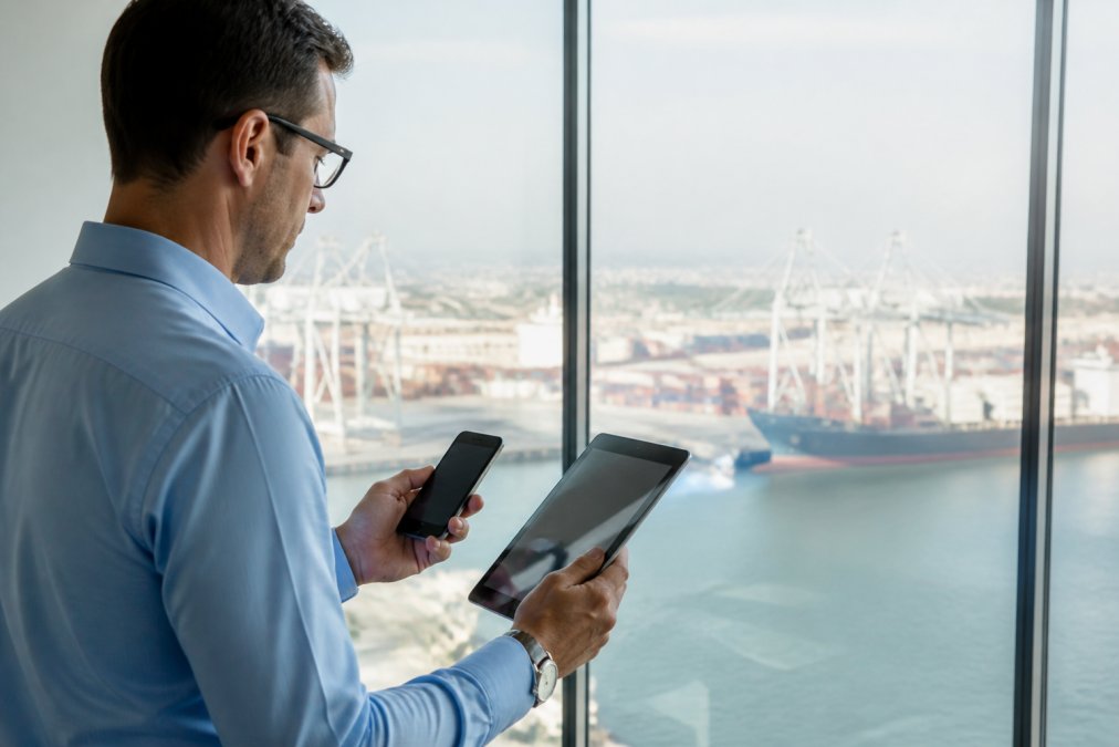 Smartphone and tablet displaying real-time vessel tracking interface with port terminals, container information, and live location data visible on screens; professional setting with natural light