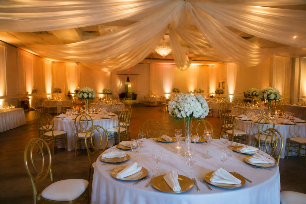 Elegant banquet hall set for a formal event with draped white fabric ceiling, round tables with gold chargers, and warm up-lighting on the walls