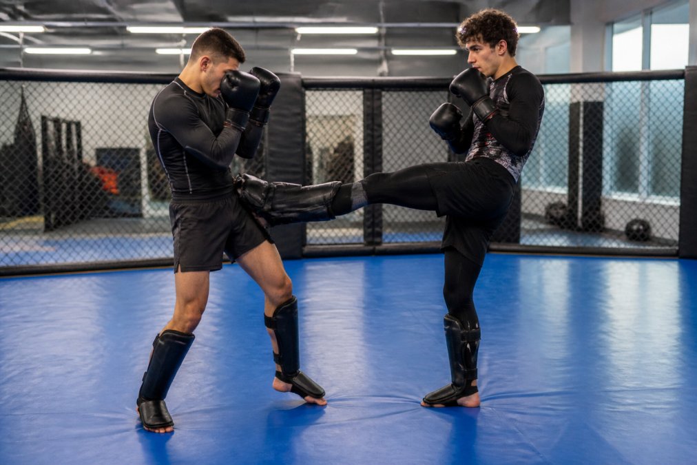 Twee beginnende MMA-sporters in een mma starterset sparren licht op een blauwe trainingsmat in een verlichte sportschool.