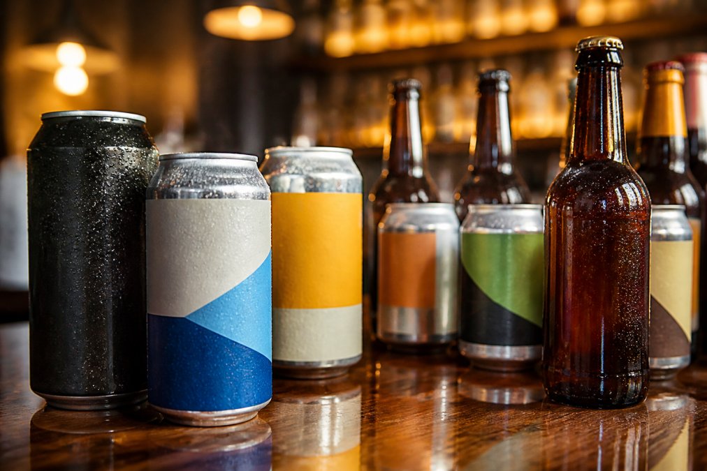 Close-up of craft alcohol-free beer cans and bottles on a wooden bar counter with condensation droplets, rich amber and dark colours visible, warm bar lighting