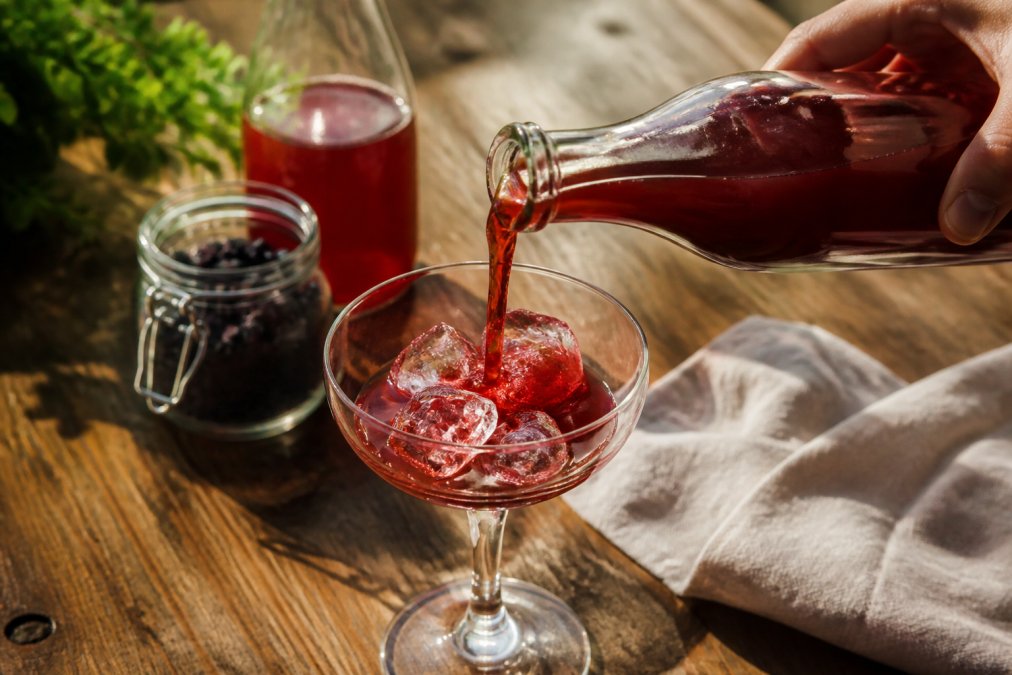 A hand pouring ruby-red shrub mixer from a glass bottle into a coupe glass filled with ice, showing the rich burgundy color and glossy texture of the fermented liquid catching warm afternoon light