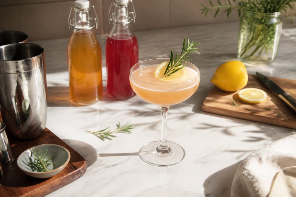 A finished mocktail in a coupe glass garnished with fresh rosemary sprig and thin lemon wheel, surrounded by bottles of raw kombucha and shrub mixer on a marble countertop with warm natural light creating shadows